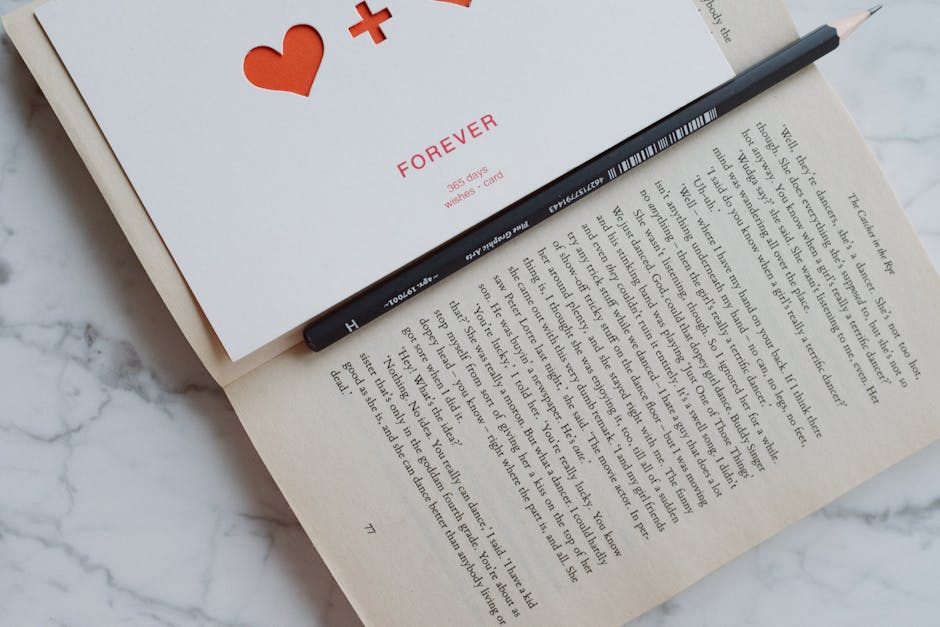 Top view of opened book with romantic postcard and black pencil composed on white marble table in modern apartment
