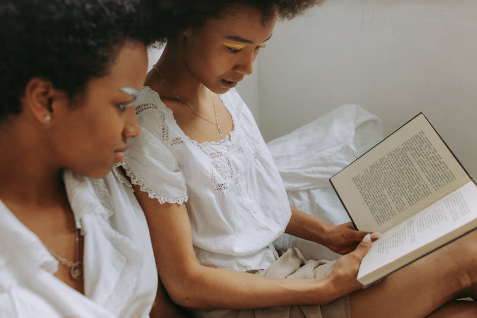 Two women with afro hair reading a book together in a cozy indoor setting, both wearing white dresses.