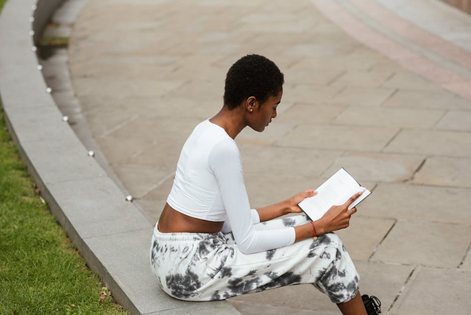 From above side view of young concentrated ethnic female with Afro haircut reading book on urban pavement