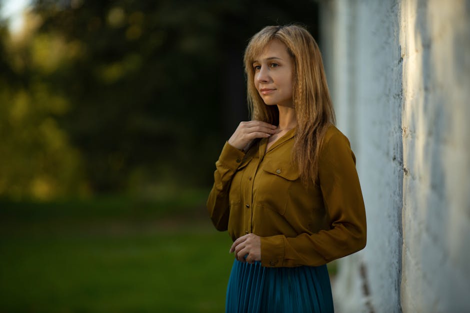 Portrait of a woman wearing a mustard blouse standing by a white wall in a natural setting.
