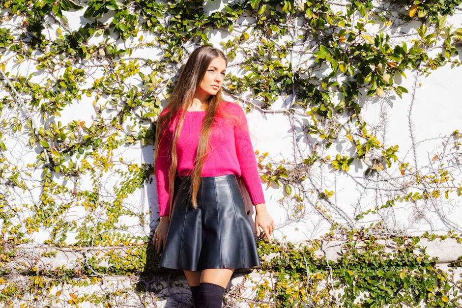 Woman in pink sweater and black skirt posing by ivy-covered wall in sunlight.
