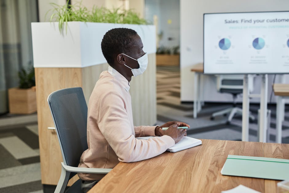 A man wearing a face mask sits at a desk in a modern office setting.