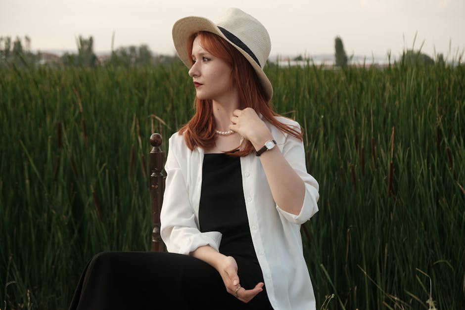 A fashionable woman wearing a sunhat and white shirt poses elegantly in a lush summer field.
