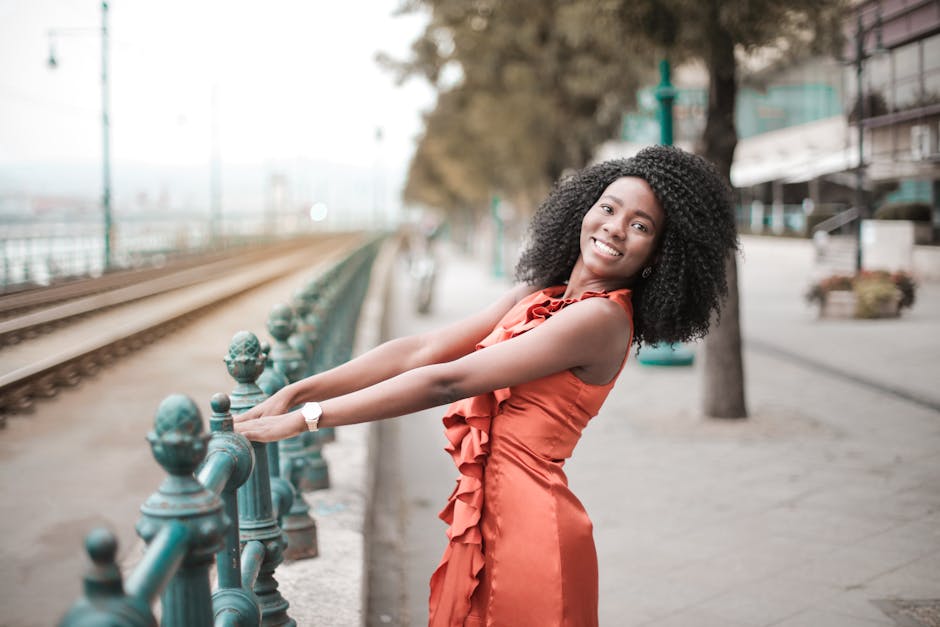 A cheerful woman in an orange dress poses by a metal railing on a city street.